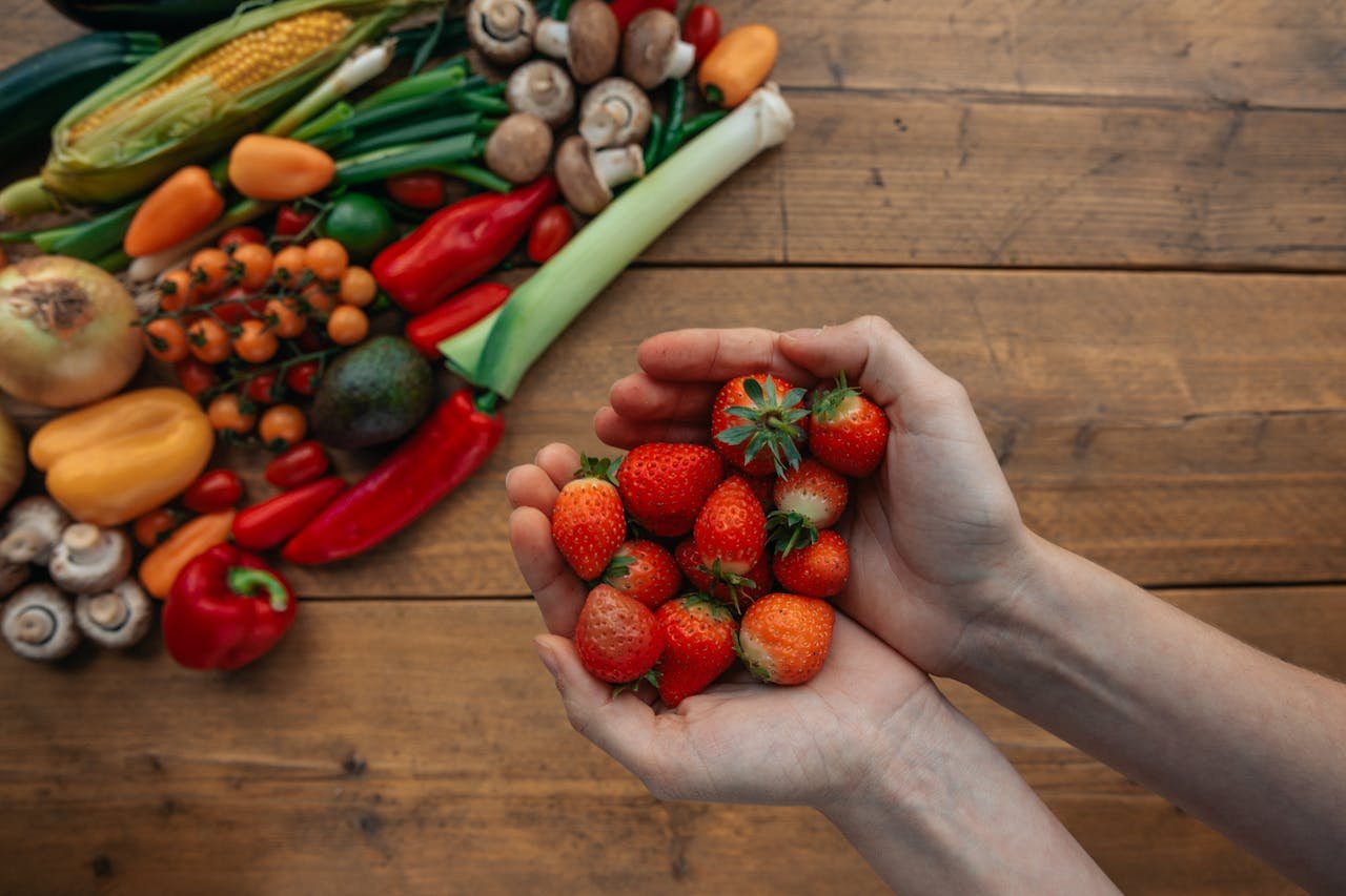 Hands holding fresh strawberries above a variety of colorful vegetables on a wooden table.